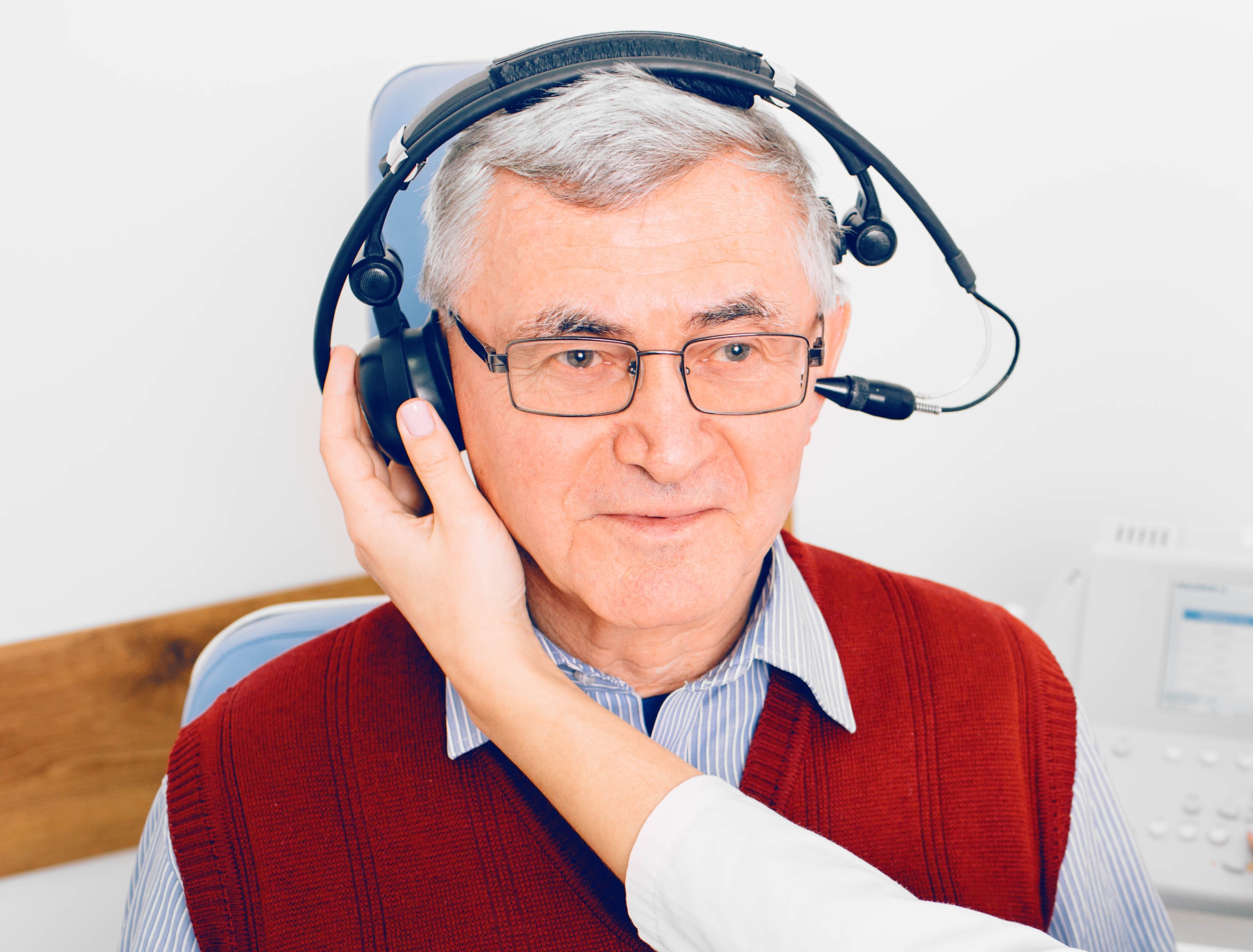 Senior man draagt koptelefoon, hij zit naast audicien tijdens een hoortest in een kliniek