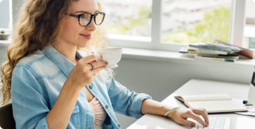 jonge vrouw zit achter haar computer met een kopje koffie in haar hand
