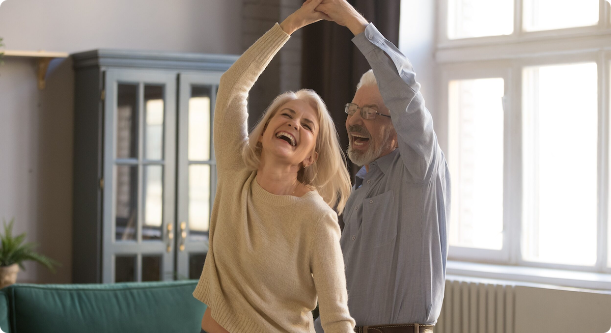 Overgelukkige volwassen grijsharige blanke man en vrouw hebben plezier en genieten samen van de tijd thuis, gelukkige bejaarde echtgenoten dansen in de woonkamer, senior man zwaait glimlachende vrouw van middelbare leeftijd