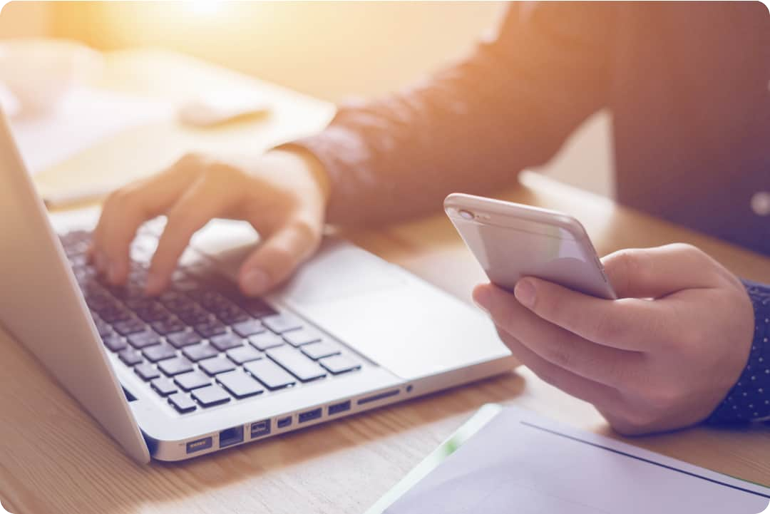 Man zit aan een bureau met zijn ene hand op het toetsenbord van zijn laptop en in zijn andere hand een smartphone