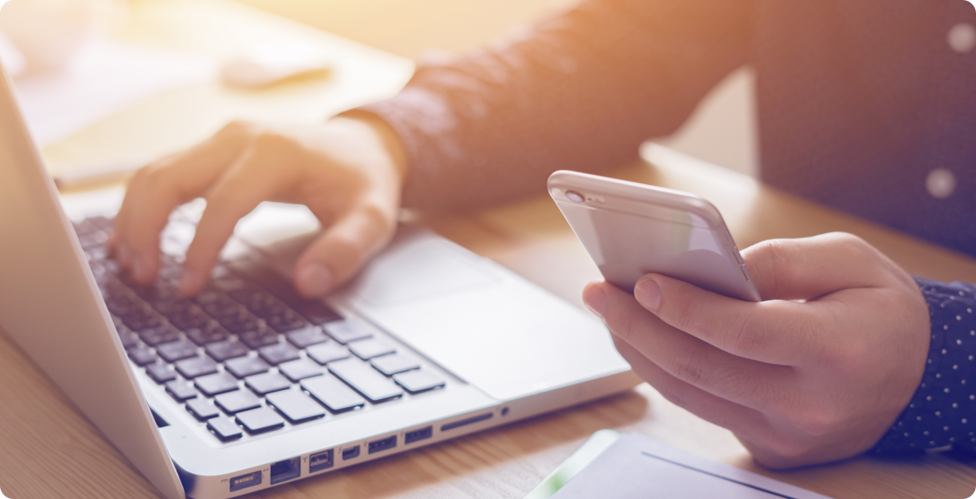 man zit achter bureau met een laptop voor hem en een mobiele telefoon in zijn hand